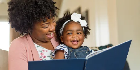 Photo: Mother reading to a smiling toddler who is sitting on her lap.