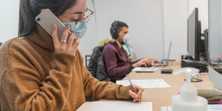 Photo: Woman working at computer desk and talking on mobile phone through a protective face mask.