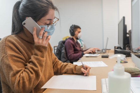 Photo: Woman working at computer desk and talking on mobile phone through a protective face mask.