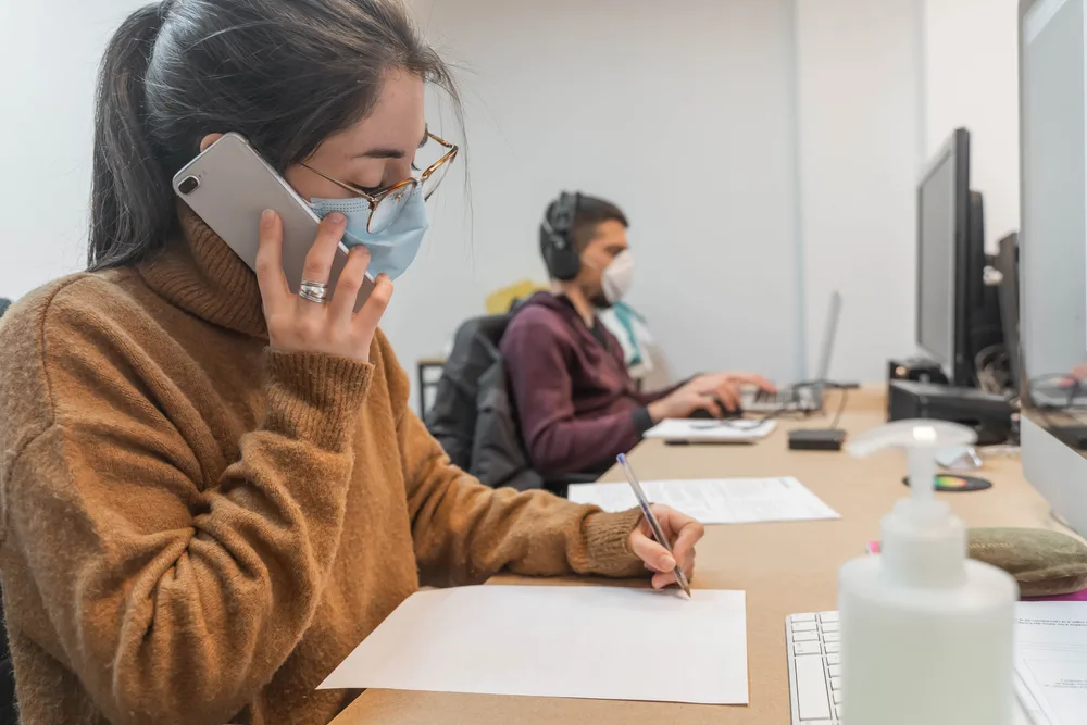 Photo: Woman working at computer desk and talking on mobile phone through a protective face mask.