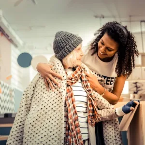 Photo: Homeless shelter volunteer smiling and putting warm coat on older homeless woman.