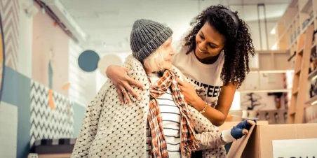 Photo: Homeless shelter volunteer smiling and putting warm coat on older homeless woman.