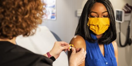 Photo: a health care provider and patient wearing a face mask, inside a clinical setting, the provider is placing a colorful bandage of the vaccination shot site on the patient's upper arm. (Source: CDC, unsplash)