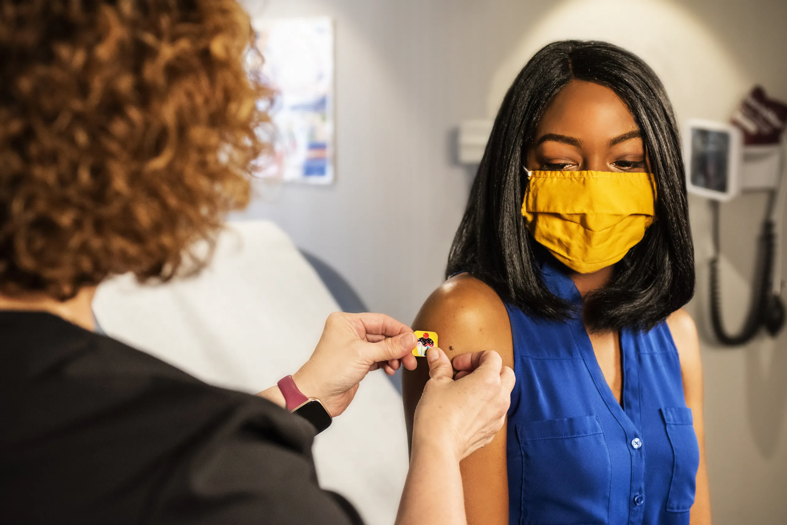 Photo: a health care provider and patient wearing a face mask, inside a clinical setting, the provider is placing a colorful bandage of the vaccination shot site on the patient's upper arm. (Source: CDC, unsplash)