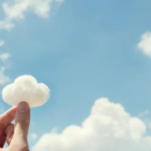 Photo: Two fingers pinched together to hold a tuft of cotton shaped like a cloud with blue sky and real clouds in the background.