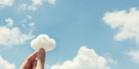 Photo: Two fingers pinched together to hold a tuft of cotton shaped like a cloud with blue sky and real clouds in the background.