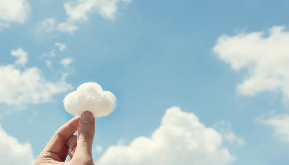 Photo: Two fingers pinched together to hold a tuft of cotton shaped like a cloud with blue sky and real clouds in the background.